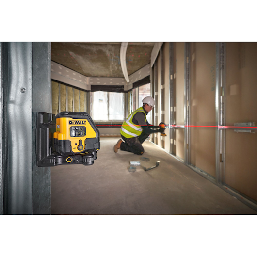 The DEWALT USB Red Beam Cross Line Laser angled in foreground attached to metal frame using the magnetic wall bracket, red laser is on and worker is onsite in the background.