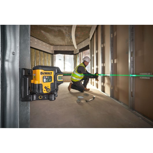 The DEWALT USB Cross Line +5 Spot Green Beam Laser angled in foreground attached to metal frame using the magnetic wall bracket, green laser is on and worker is onsite in the background.