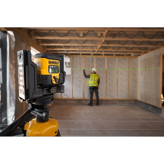 The DEWALT 3x 180 USB Green Beam Laser in foreground attached to tripod, green laser is on and worker is onsite in the background.