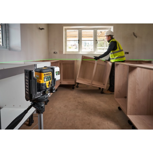 The DEWALT 3x 180 USB Green Beam Laser in foreground attached to tripod, helping to guide the worker on site fitting cabinets.