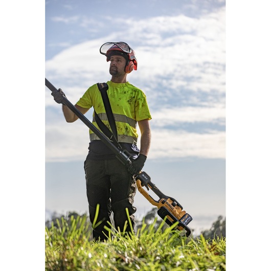 Worker in high visibility top and helmet with visor up and safety harness getting ready to use the extended DEWALT 54V Tele Pole Saw 4.0M 30CM on tree in open large field.