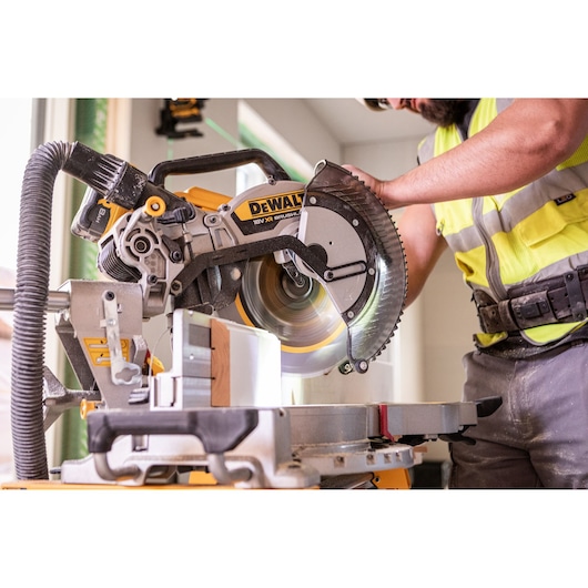 The DEWALT 18V XR 305mm Slide Mitre Saw being used on job site by worker in high visibility vest, safety goggles and helmet, worker is cutting through wooden beam.
