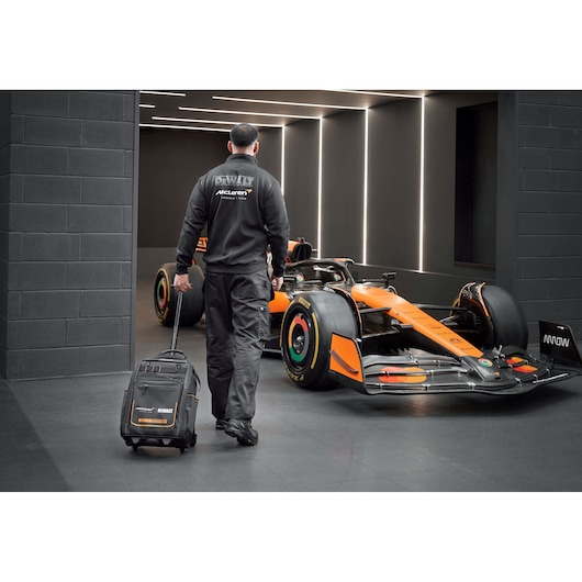 DEWALT/McLaren backpack on wheels being pulled by a McLaren mechanic walking towards a McLaren racing car