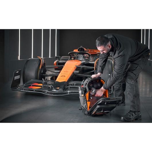 McLaren mechanic placing a drill driver into a DEWALT/McLaren backpack on wheels. A McLaren racing car is in the background