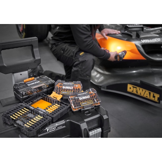 x4 Limited Edition DEWALT/McLaren screwdriver bit sets sit on top of a Tstak box. In the background a mechanic is working on a McLaren racing car.