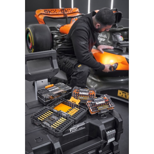 x4 Limited Edition DEWALT/McLaren screwdriver bit sets sit on top of a Tstak box. In the background a mechanic is working on a McLaren racing car.