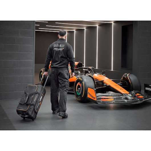 DEWALT/McLaren Duffle Bag with wheels being pulled by a McLaren mechanic walking towards a McLaren racing car
